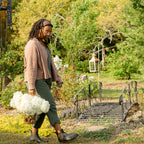 Quilenthia walking through a garden with a bouquet of flowers in the Linden Glass & Wicker Vase , surrounded by greenery and a small bridge.