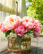 Pink peonies in a glass vase with wicker base on a wooden surface outdoors.