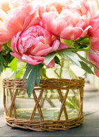 Pink peonies in a glass vase with wicker insert base on a wooden surface