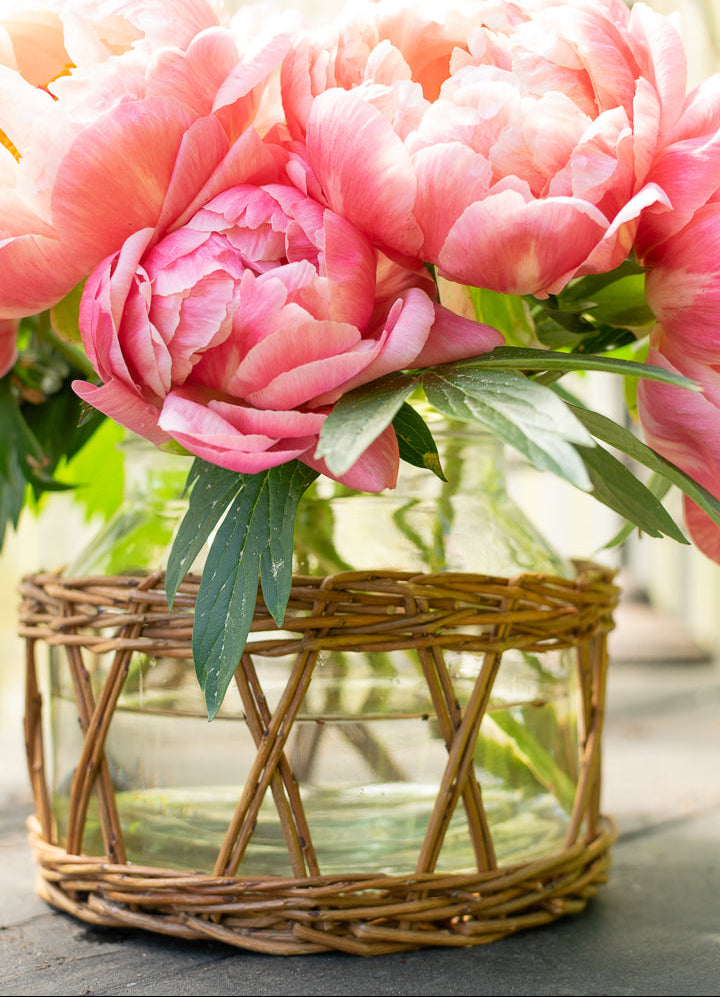 Pink peonies in a glass vase with wicker insert base on a wooden surface